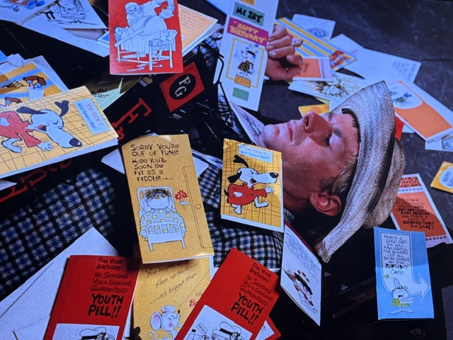 A person lying on the ground surrounded by colorful cartoon postcards featuring humorous messages and illustrations, including characters like Snoopy.