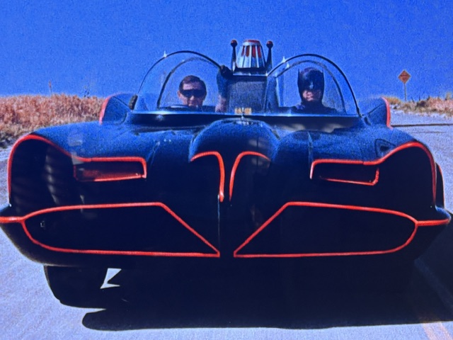 A classic black batmobile with red outlines on a road, featuring two characters inside, one resembling Batman.