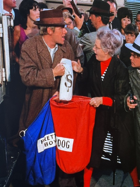 A man in a brown coat and hat is holding a white bag marked with a dollar sign, while a woman with gray hair in a red shirt and black jacket holds a red bag labeled 'DOG'. They are surrounded by a crowd in an urban setting.