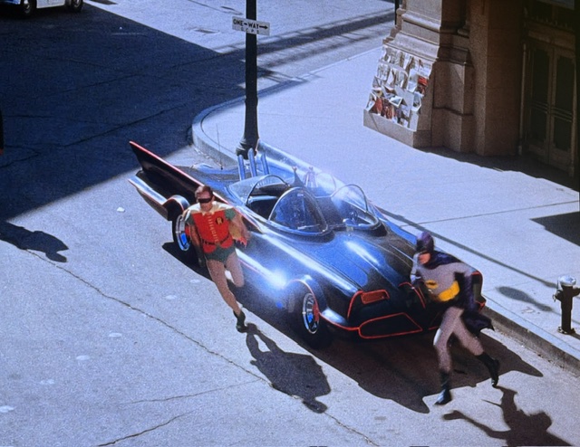 Batman and Robin running towards the Batmobile in a city street.