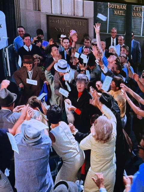 A large crowd gathered outside a bank, with people enthusiastically throwing papers and raising their hands. The scene captures a sense of excitement and chaos in front of the Gotham City National Bank.
