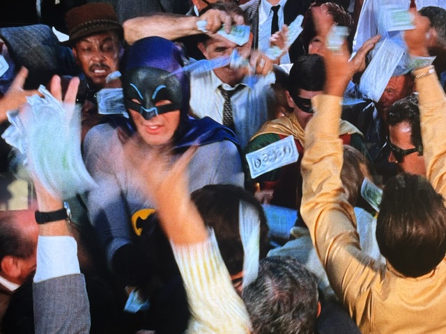 A chaotic scene featuring a crowd of people reaching for money while two figures in superhero costumes, one resembling Batman, are central to the commotion.