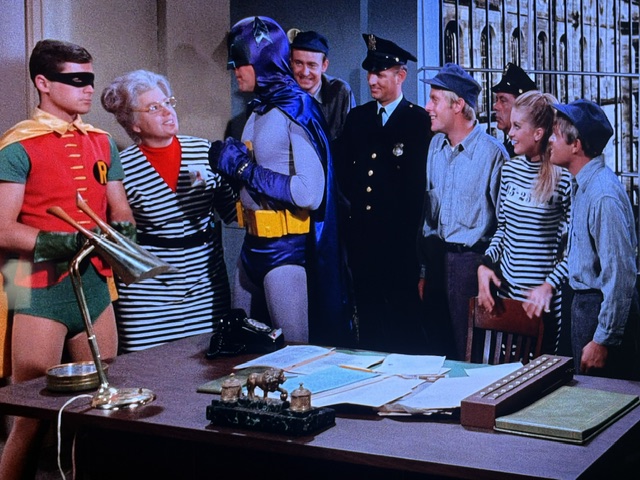 Batman and Robin interacting with a group of characters in a police station setting, featuring various outfits including striped prisoner uniforms and police attire.