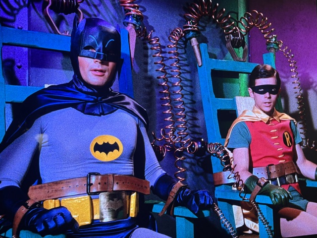 Batman and Robin seated in a brightly colored room, with them connected to futuristic devices featuring spirals and wires.