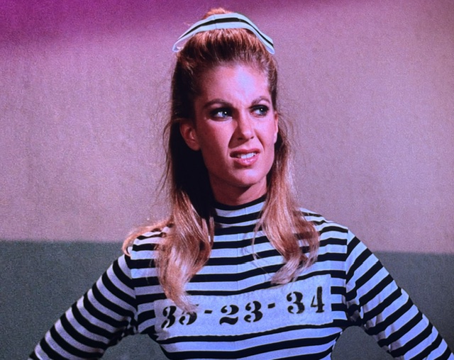 A woman with long hair wearing a black and white striped jail outfit, looking displeased, with a headband in her hair.