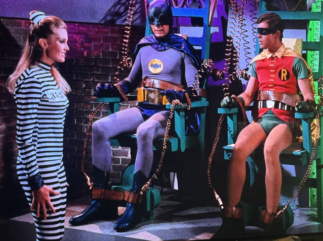 A scene from a classic superhero show featuring a female character in a striped prison outfit standing in front of Batman and Robin, who are sitting in high-tech chairs restrained with colorful wires.