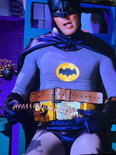 A character dressed as Batman, wearing a blue and gray costume with a yellow bat symbol and a utility belt, sitting in a chair with an intense expression.