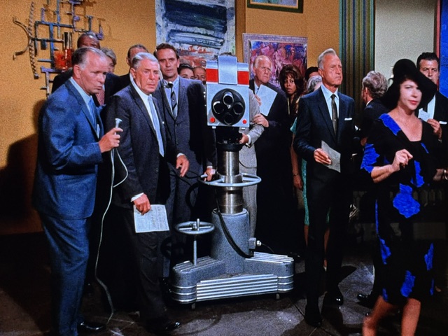 A group of people, dressed in formal attire, gathered around an old-fashioned television camera on a stand. One man is speaking into a microphone, while others hold papers and observe. A woman in a blue dress is seen moving away from the group.