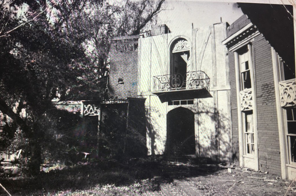 Black and white photo of a partially collapsed building surrounded by trees, featuring an open balcony and ornate architectural details.