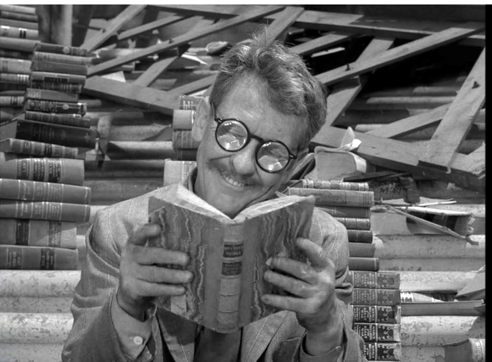 A smiling man wearing glasses is sitting among stacks of books, reading a book with enthusiasm.