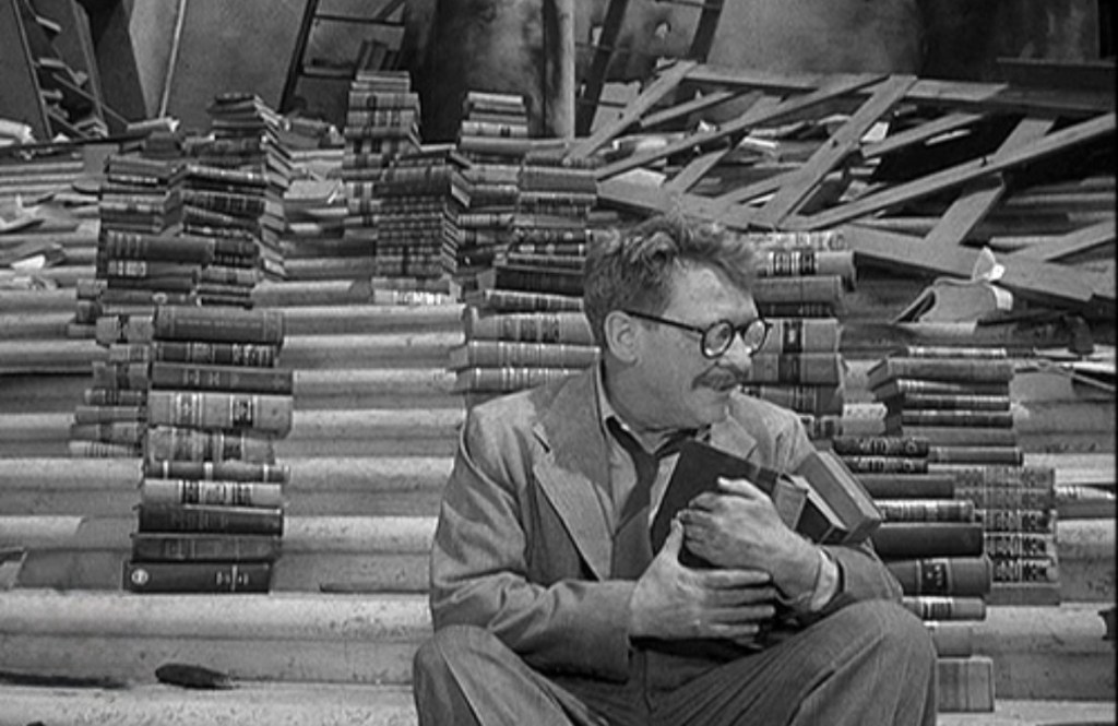 A man sitting on stairs surrounded by stacks of books, holding a few close to his chest, with a distressed expression amidst a chaotic backdrop.