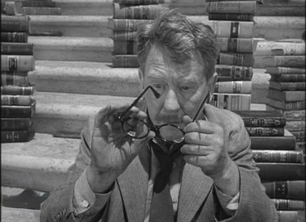 A man with gray hair examines a pair of glasses while surrounded by stacks of books on steps.