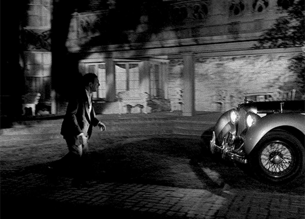 A man in formal attire walks towards a vintage car parked in front of a large, shadowy building at night.