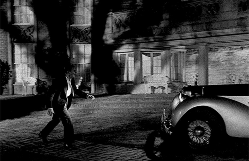 A man in formal attire walks towards a vintage car in front of a large, shadowy house at night.