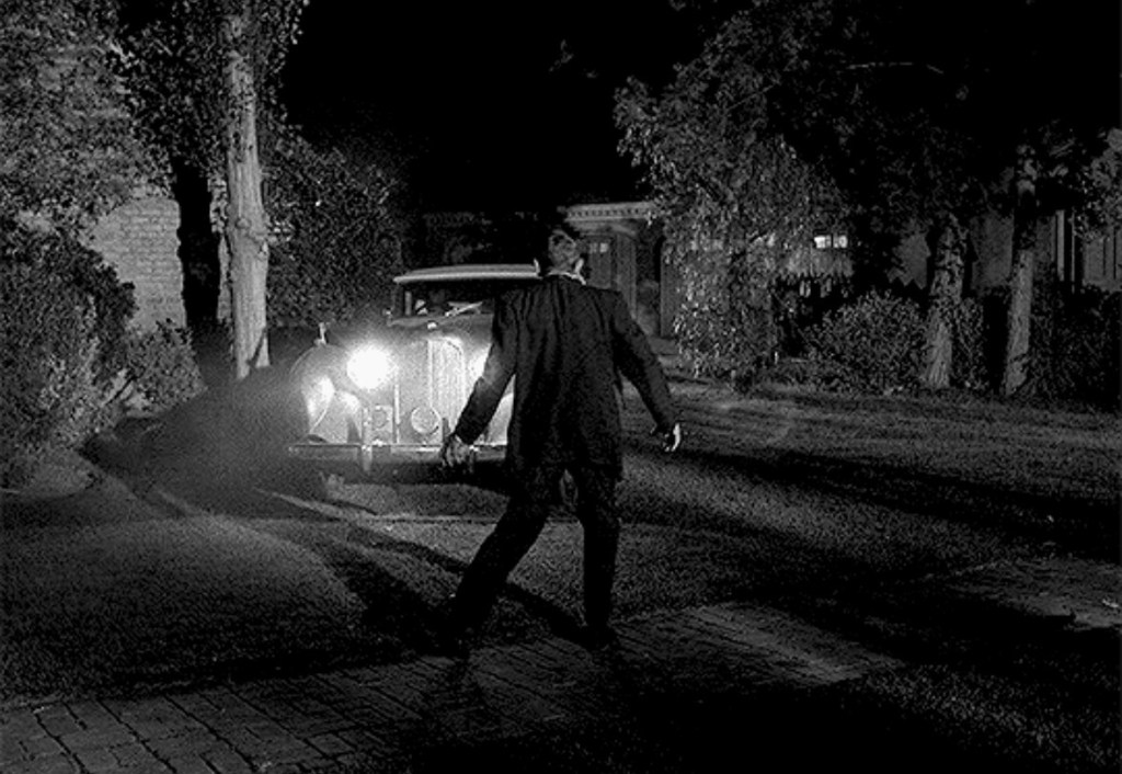 A man stands in front of a vintage car with headlights shining, set in a shadowy outdoor environment at night.