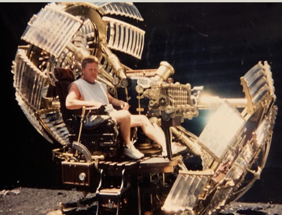 A person seated in a mechanical device resembling a futuristic machine with large, intricate components and transparent panels.
