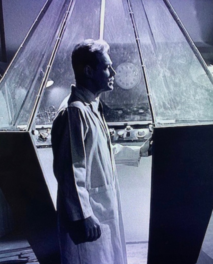 A man in a lab coat stands beside a large, angular scientific device with a transparent top, surrounded by various dials and instruments, depicted in black and white.