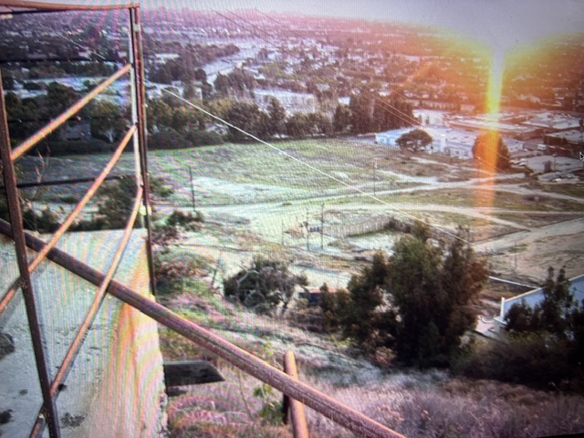 View from a hillside overlooking a landscape with a sunset, featuring a metal railing in the foreground and distant structures below.