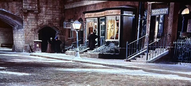 A snowy city street scene featuring a storefront labeled 'Wylie's Dainty Store,' with shop windows displaying items. People dressed in period clothing are gathered outside the store, and a lamp post illuminates the cobblestone path. The architecture reflects a historical style, with brick buildings and a staircase leading to a nearby structure.