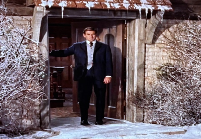 A man in formal attire stands at the entrance of a house, surrounded by a snowy landscape and icicles hanging from the roof.