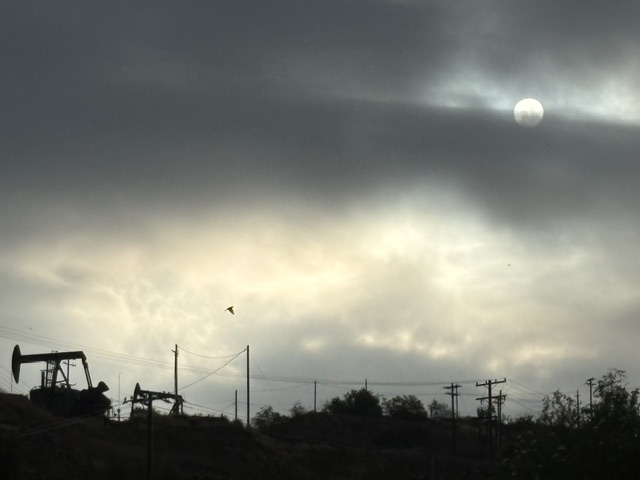 Silhouette of an oil pump jack against a cloudy sky with the sun partially obscured.