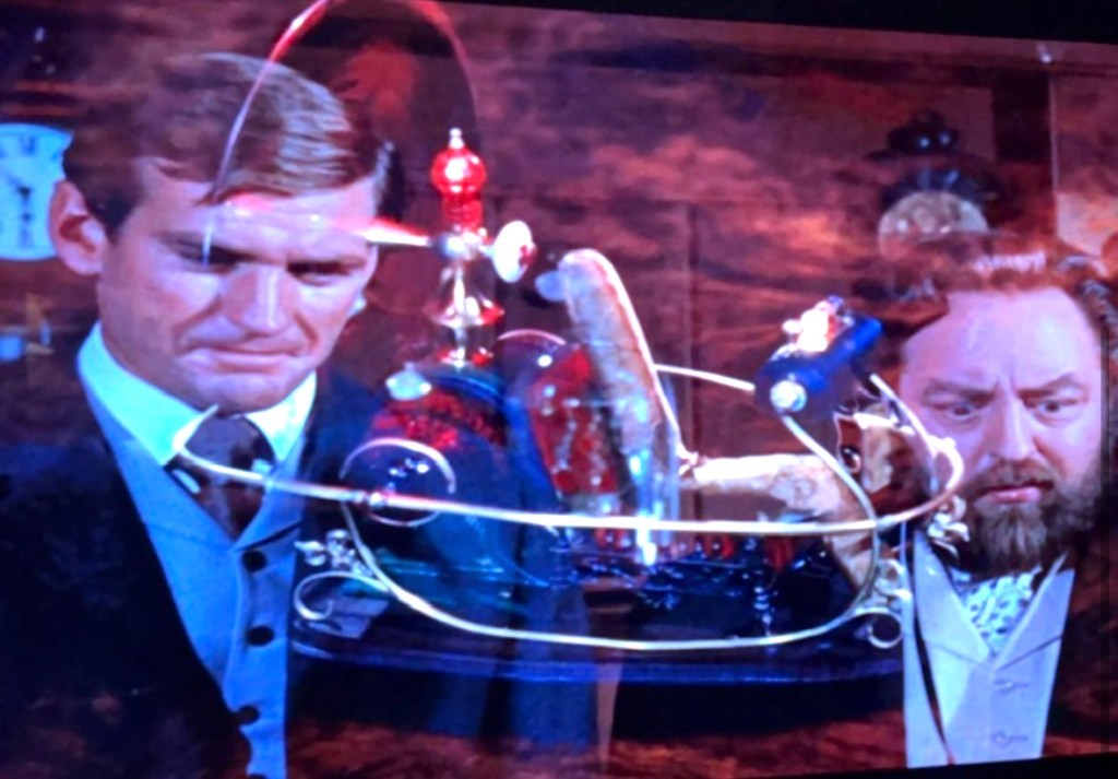Two men in vintage clothing examining a complex mechanical device with colorful components in a dimly lit room.