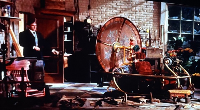 A vintage workshop interior featuring a large, whimsical time machine with colorful details and a prominent wheel, alongside various tools and items scattered around.