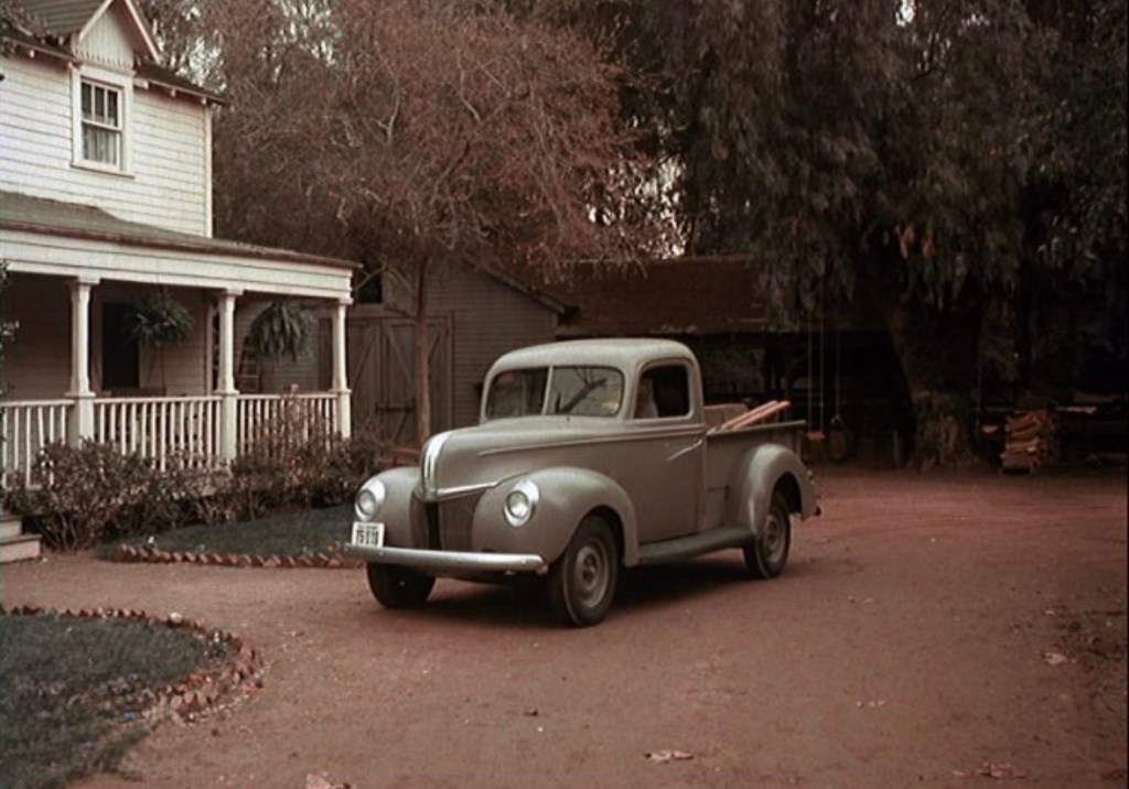 A vintage gray pickup truck parked in front of a house with a porch and greenery, set in a rural environment.