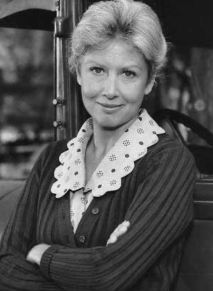 A woman with short hair, wearing a cardigan and a polka-dotted collar, smiles confidently while standing with her arms crossed, in front of a vintage vehicle.