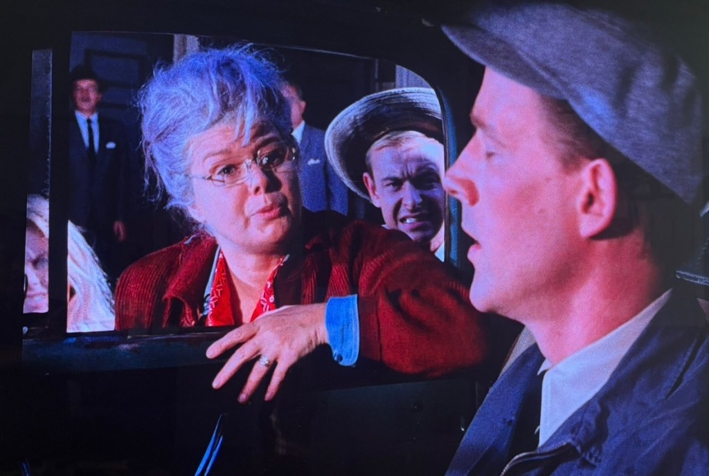 A scene featuring a woman with gray hair and glasses speaking to a man in a cap inside a car, while a man in the background looks on with a curious expression.