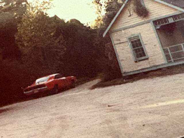 A vintage red car parked near a small, old house with a porch in a rural setting surrounded by trees.