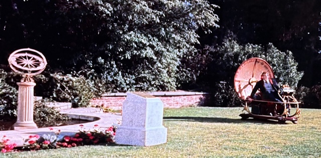 A man sitting in a futuristic, wheeled device in a garden setting, with a stone monument and a decorative structure nearby, surrounded by greenery and flowers.