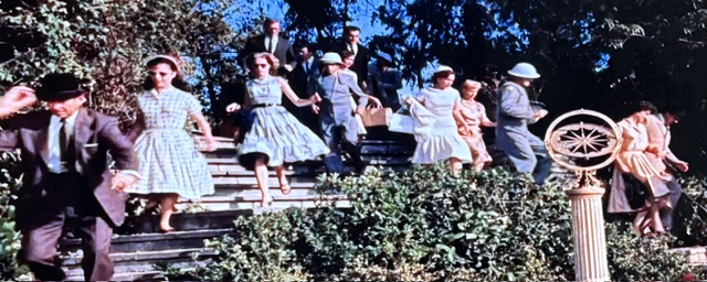 A group of people, including children and adults, walking down steps surrounded by greenery on a sunny day.