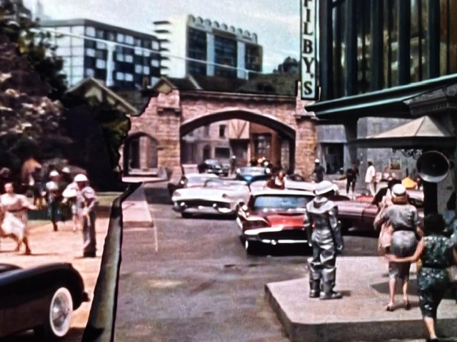 A busy urban street scene featuring vintage cars, pedestrians, and a historic archway. The foreground shows people walking, with a shop sign visible in the background.