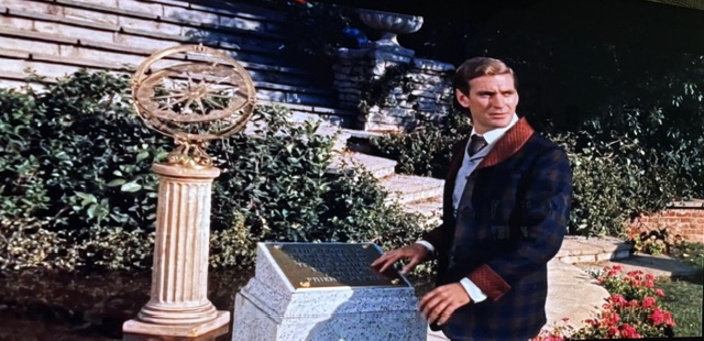 A man in a formal outfit stands near a decorative compass on a pedestal, with a stone plaque beside him, set in a lush garden.