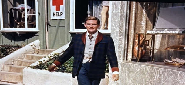 A man in a stylish outfit, consisting of a plaid jacket and vest, walking confidently in front of a storefront with a red cross sign and a mannequin in the window.