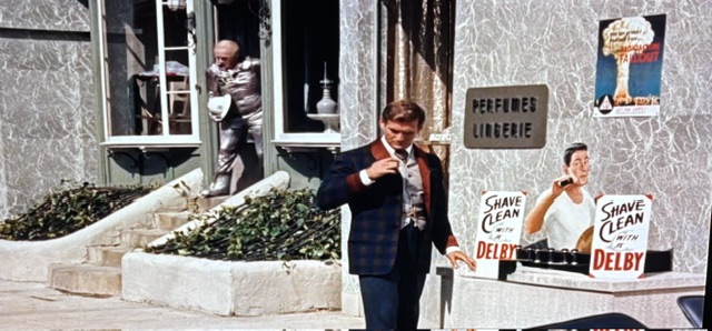 A man in formal attire stands in front of a building with a sign for 'Perfumes Lingerie,' holding a drink and looking at a poster for 'Shave Clean with Delby.' In the background, another man dressed in silver is exiting the building.