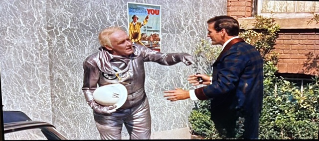 A man in a shiny silver suit holding a round object is gesturing animatedly while talking to another man in a dark blazer. The background features a vintage poster on a textured wall.