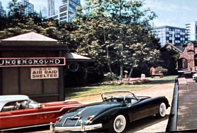 A vintage scene featuring two classic cars parked near an underground air raid shelter sign, with greenery and skyscrapers in the background.