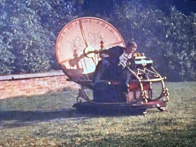 A man operating a whimsical time machine with a large clock face and intricate mechanisms, set against a grassy landscape.
