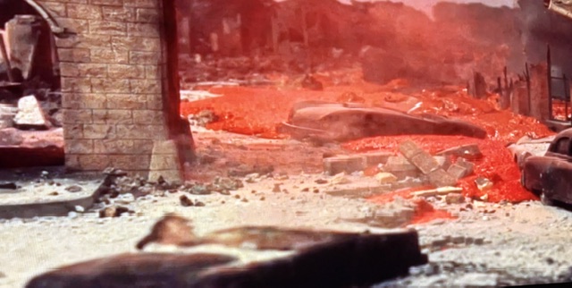 A devastated landscape featuring ruined buildings, debris scattered on the ground, and bright red liquid resembling lava flowing through the scene.