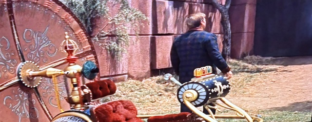A man wearing a checkered jacket stands with his back to the camera, looking at a large ornate door. Beside him is a colorful, vintage-style time machine with red upholstery and various decorative elements.