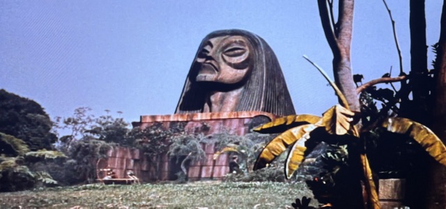 Large, abstract stone statue resembling a head, surrounded by lush greenery and tropical plants.