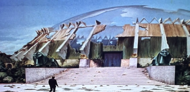 A lone figure stands before a large, dilapidated structure with a dome-shaped roof, featuring broken elements and overgrown surroundings.
