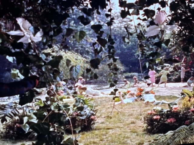 A view through lush greenery overlooking a tranquil scene by a river, with people engaging in various activities along the water's edge.