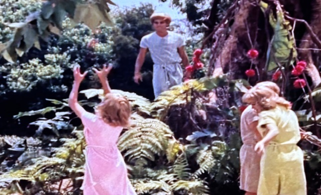 Children playing in a lush outdoor setting, with two girls in light dresses reaching out towards a boy standing nearby among tropical plants.