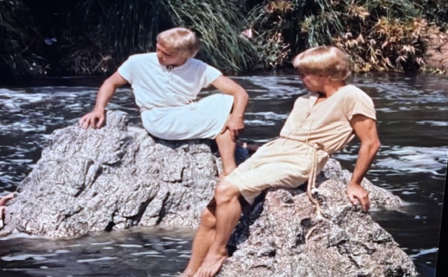 Two young men sitting on a rock by a river, wearing light-colored tunics, with water and greenery in the background.