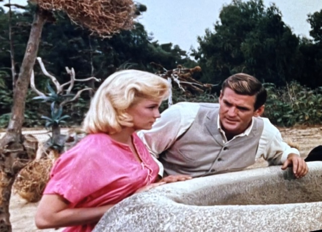 A blonde woman in a pink shirt and a man in a light-colored vest lean over a stone well in a natural outdoor setting, engaging in a serious conversation.