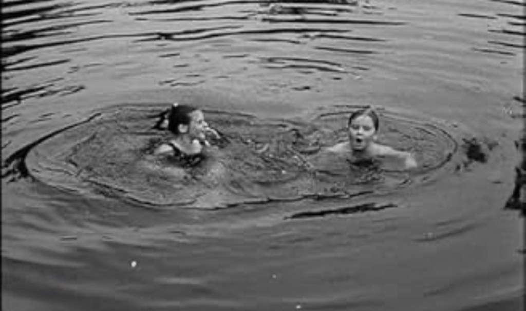 Two girls swimming in a circular formation in a lake, creating ripples on the water's surface.