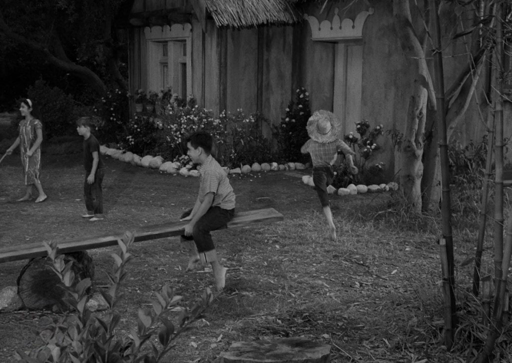 Children playing in a garden, with two boys on a seesaw and a girl holding a stick nearby, in front of a rustic house.
